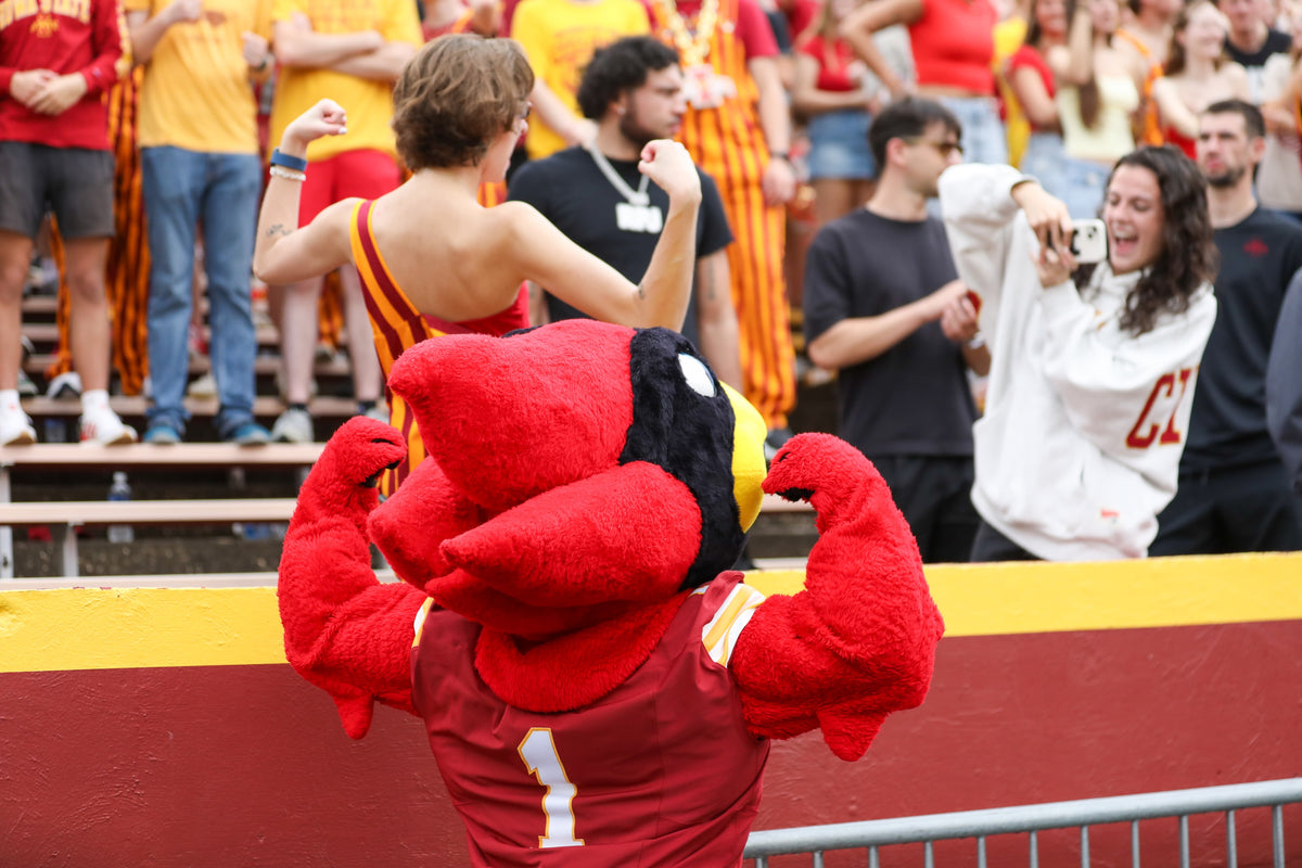 A person in a red and yellow athletic outfit poses with the Iowa State Cyclones mascot, a red character  wearing a maroon jersey with the number 1, in front of a stadium crowd.
